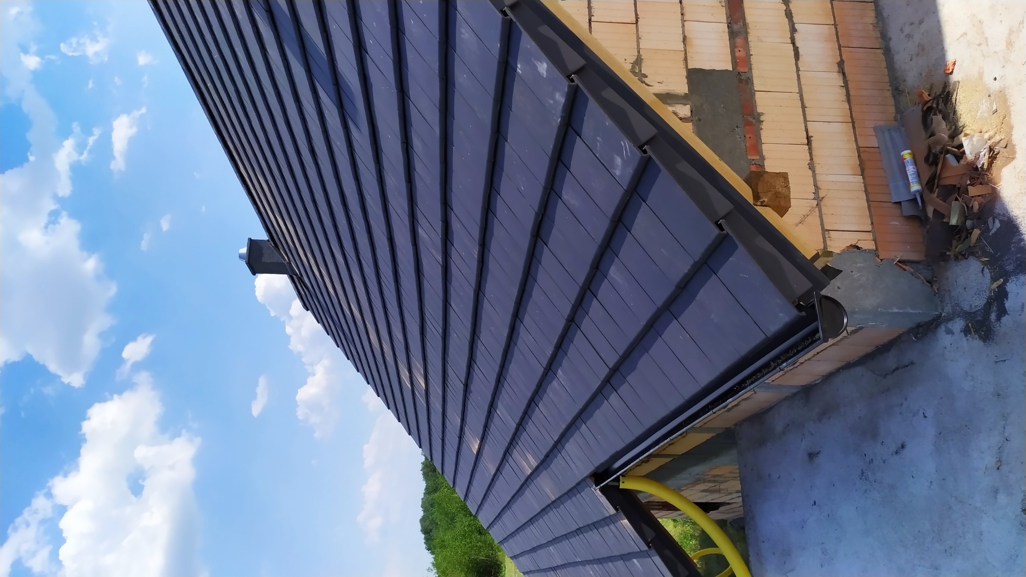 Nisko kątowe ujęcie nowo położonych, ciemnych dachówek ceramicznych z widoczną rynną i fragmentem komina na tle błękitnego nieba.