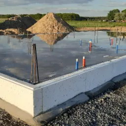 Wylany fundament z izolacją termiczną, wystające pręty zbrojeniowe i rurki instalacyjne, w tle hałdy piasku i krajobraz wiejski.