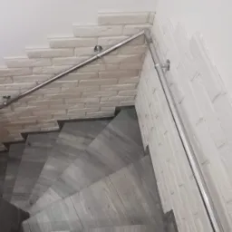 Klatka schodowa wykończona płytkami imitującymi drewno, ściany z dekoracyjnej cegły w odcieniach bieli i szarości, zabezpieczona metalową poręczą.