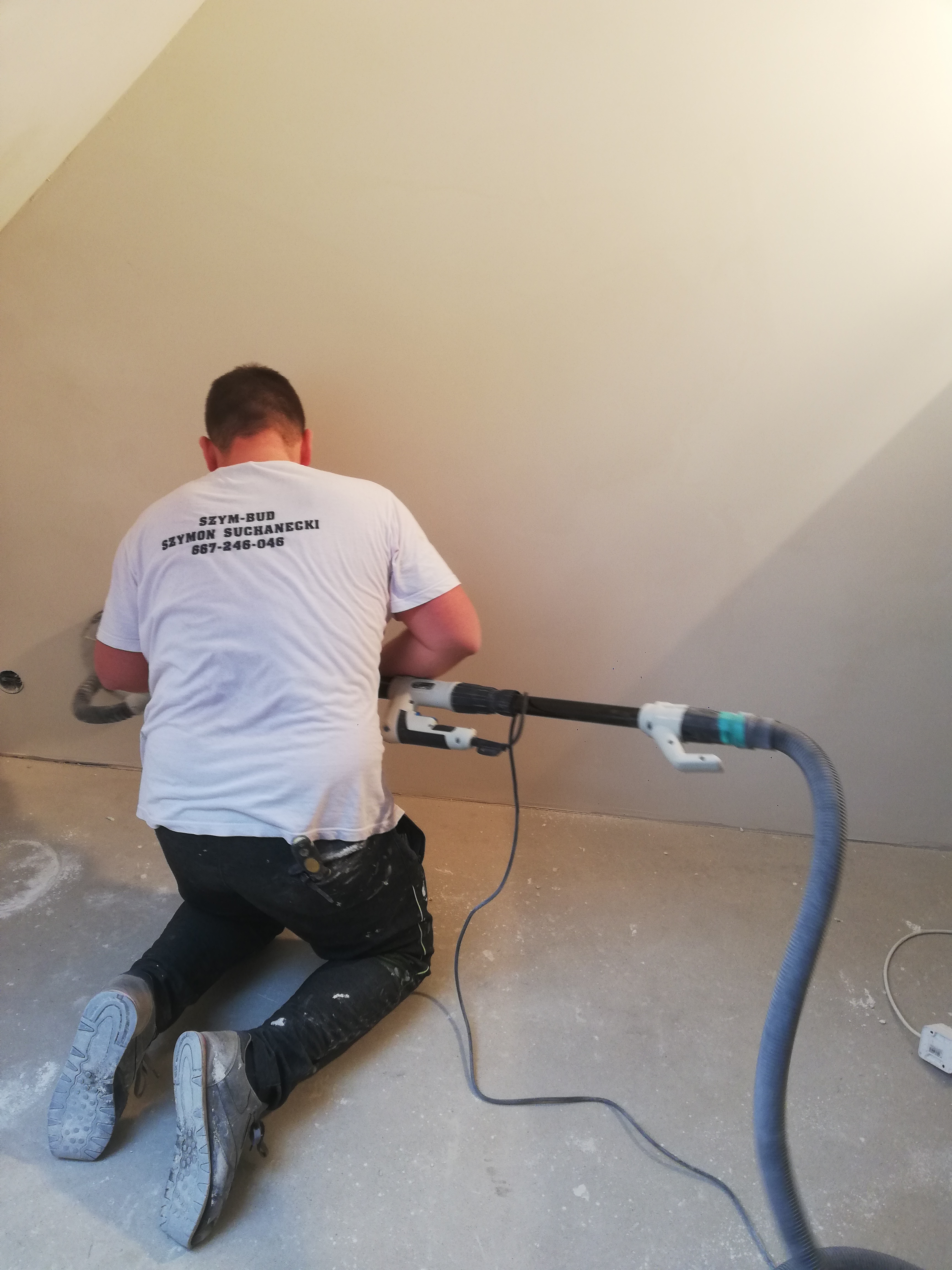 Pracownik na kolanach szlifuje ścianę gipsową szlifierką z odkurzaczem, ubrany w białą koszulkę z nadrukiem nazwy firmy 'SZYM-BUD' i numerem telefonu.