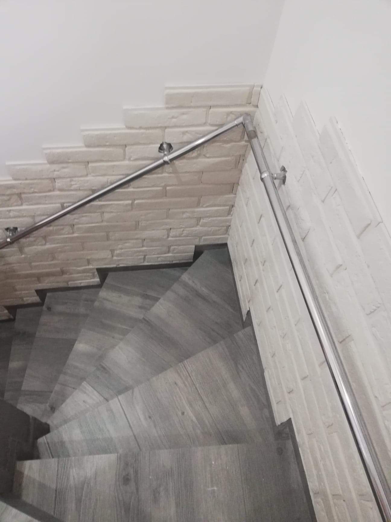 Klatka schodowa wykończona płytkami imitującymi drewno, ściany z dekoracyjnej cegły w odcieniach bieli i szarości, zabezpieczona metalową poręczą.