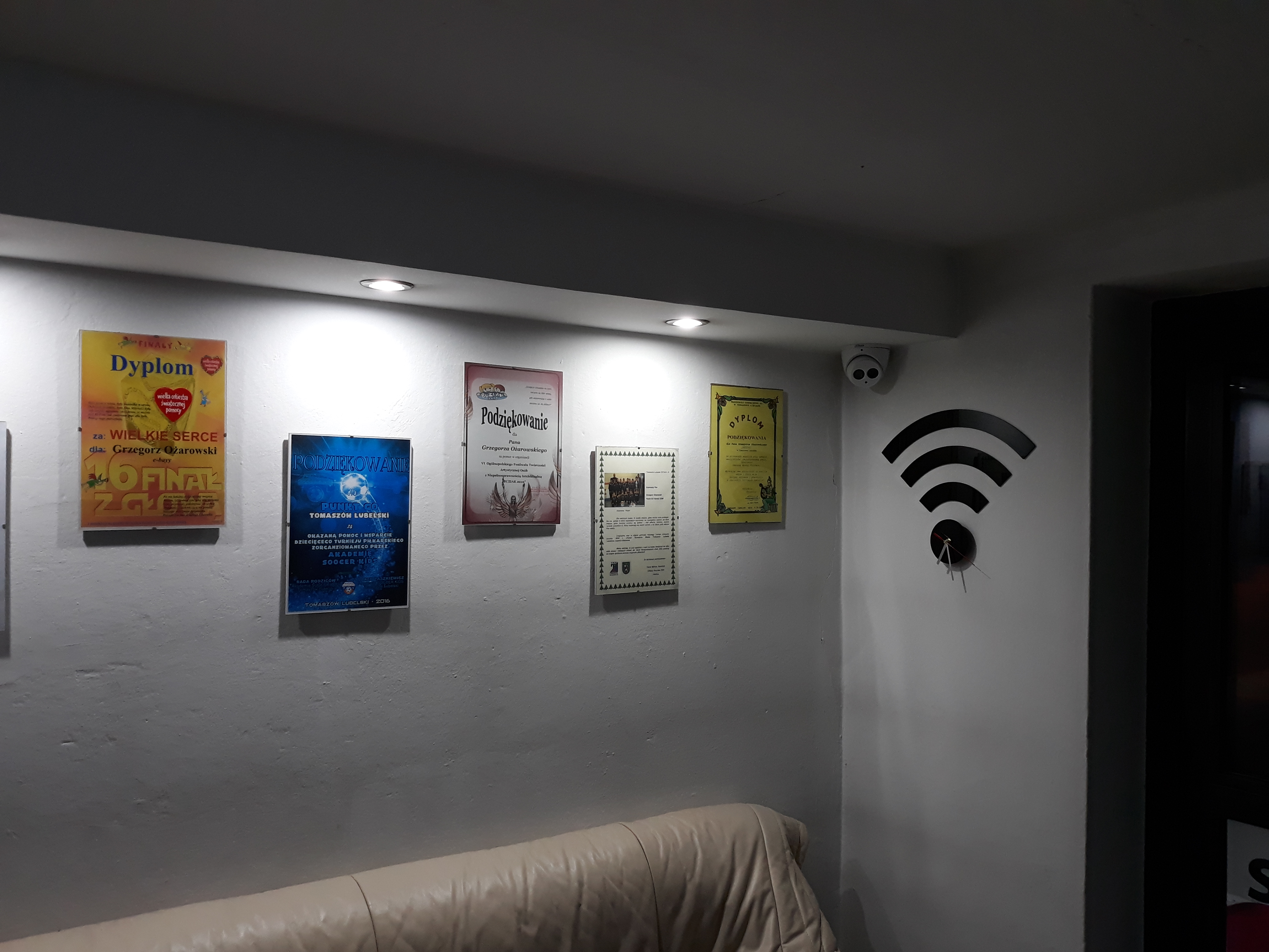Ściana w biurze lub lokalu usługowym z dyplomami, podziękowaniami i dekoracją w kształcie symbolu Wi-Fi z zegarem w środku, kamera monitoringu pod sufitem.