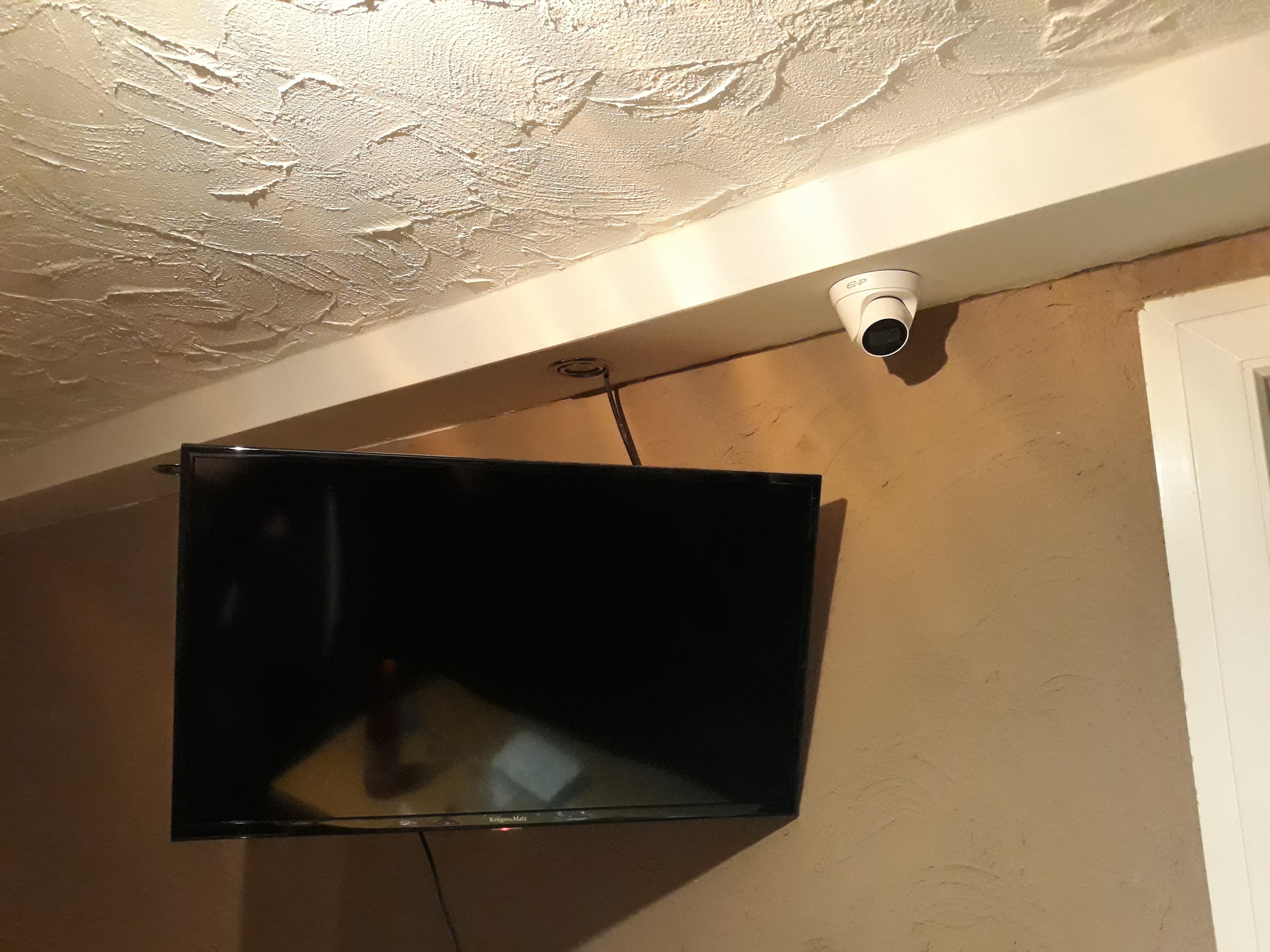 Kamera monitoringu kopułkowa zamontowana pod sufitem, obok telewizora na ścianie o nierównej fakturze. Widoczne okablowanie i fragment ramy okiennej.