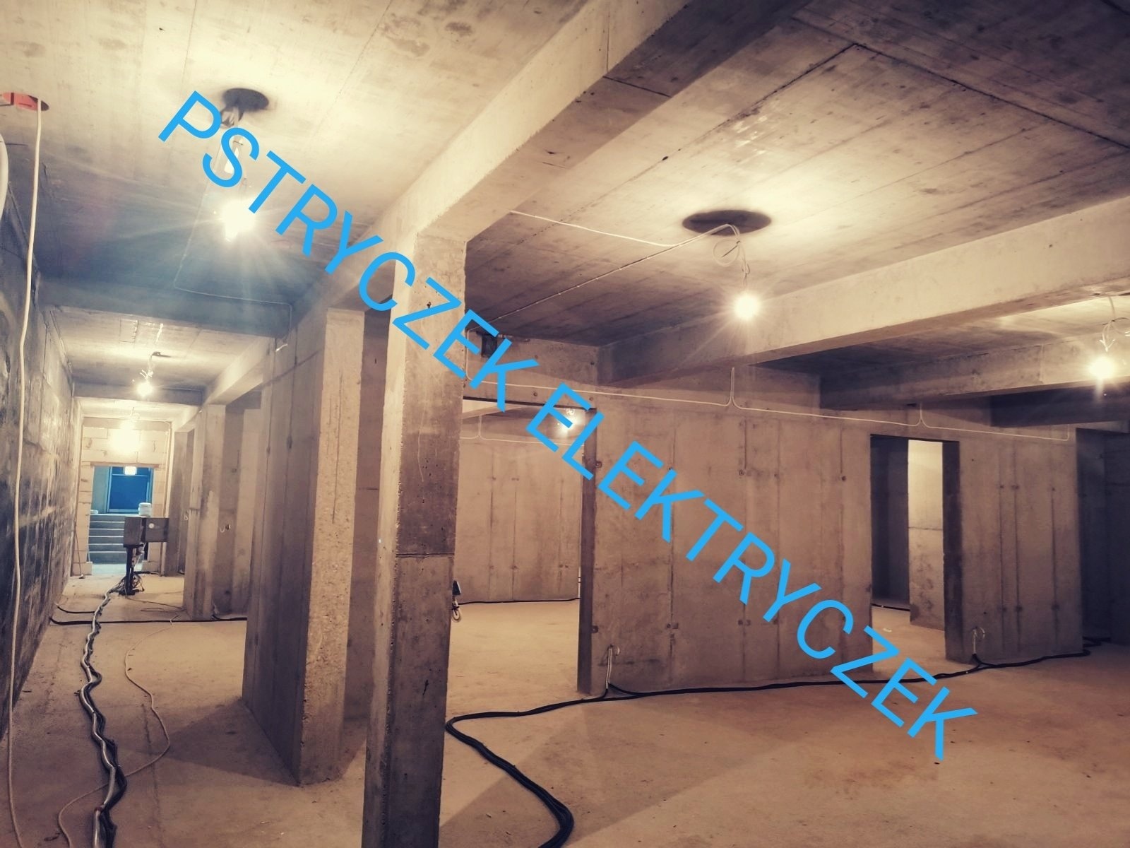 Surowy betonowy budynek w trakcie instalacji elektrycznej, widoczne kable wzdłuż ścian i żarówki pod sufitem, niskie oświetlenie.