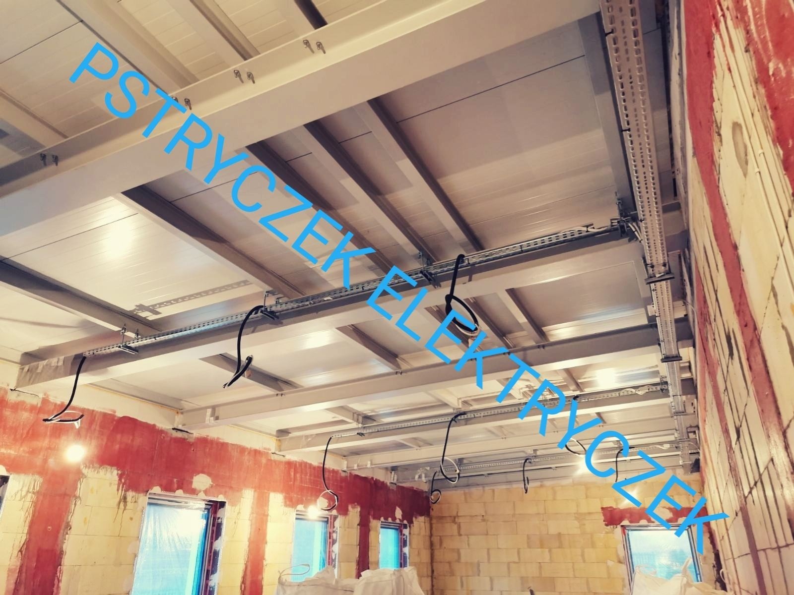 Widok z dołu na sufit i ściany w surowym stanie, z widocznymi metalowymi konstrukcjami podtrzymującymi koryta kablowe i zwisające przewody elektryczne, w tle okna zakryte folią.
