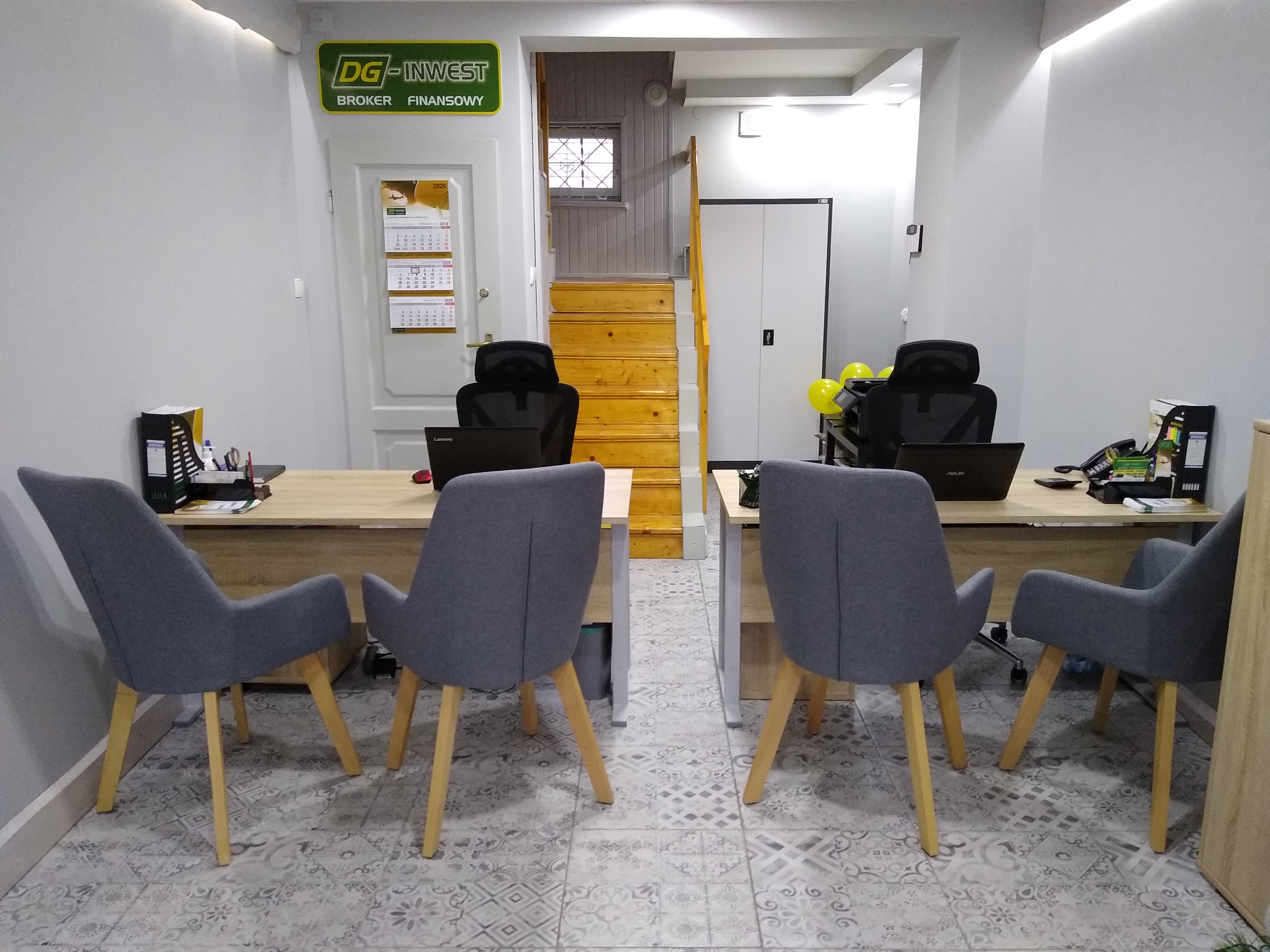 Biuro brokera finansowego z dwoma stanowiskami pracy: biurka z laptopami, szare krzesła, firmowy kalendarz na ścianie i logo DG-Invest nad drzwiami.