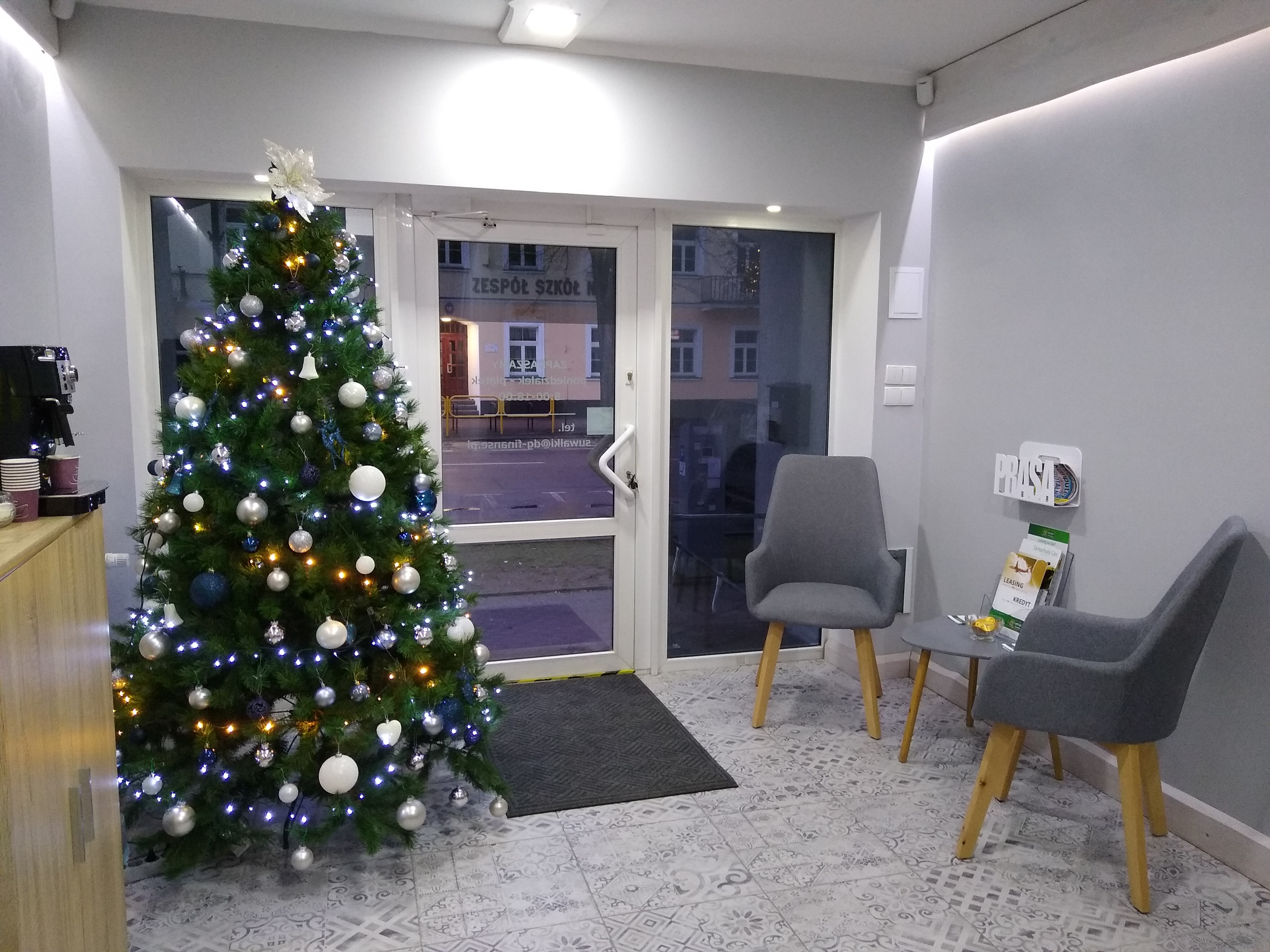 Wnętrze biura z choinką świąteczną przy wejściu, dwoma szarymi krzesłami i małym stolikiem z ulotkami informacyjnymi na temat leasingu i kredytów.