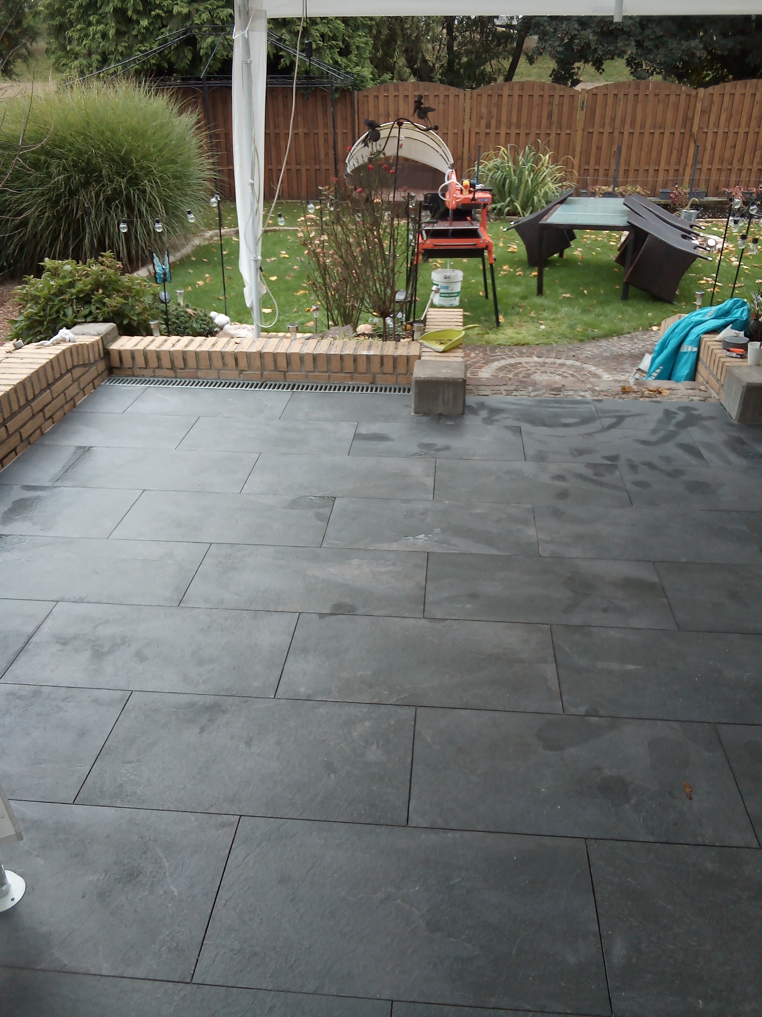 Nowo wyłożony taras z ciemnych, prostokątnych płyt, z widocznym systemem odwadniającym, w tle trawnik z elementami wyposażenia ogrodowego i narzędziami do cięcia płytek.