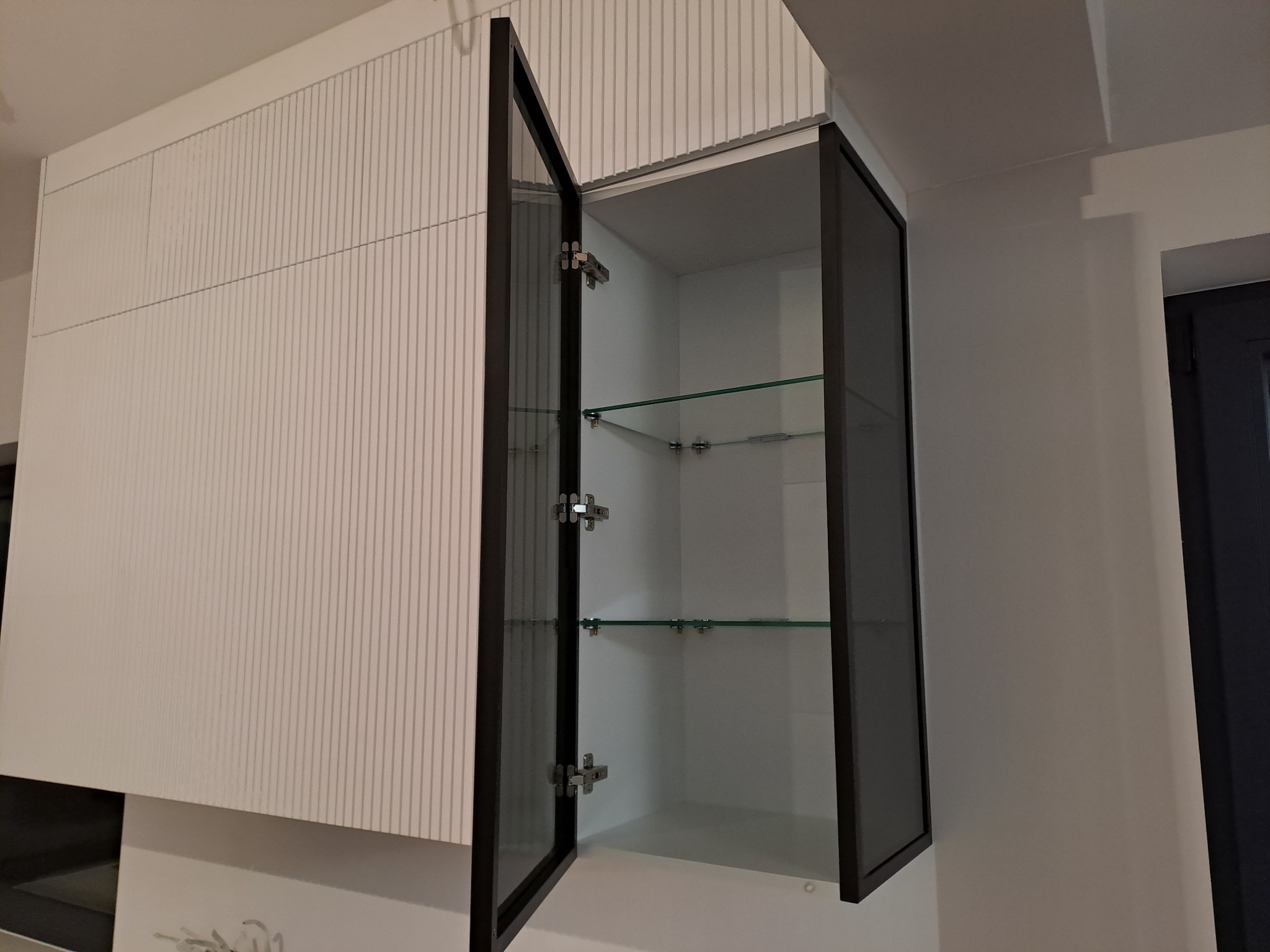 Nowoczesna szafka wisząca z białymi frontami z delikatnym pionowym wzorem i szklanymi półkami, z czarną ramą drzwi, uchylona, widoczne zawiasy.