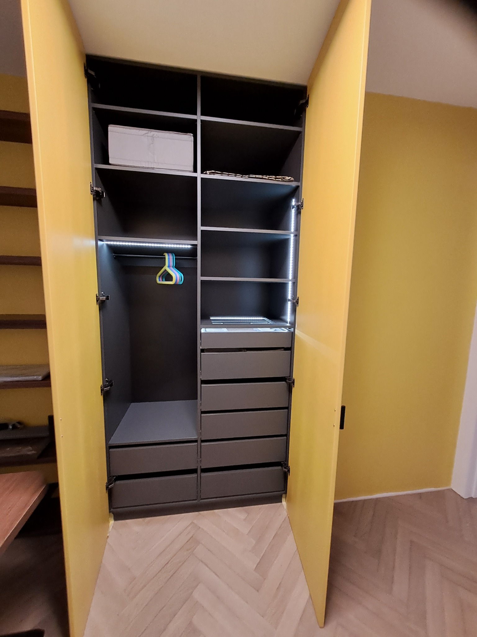 Otwarta szafa wnękowa z żółtymi drzwiami, szarym wnętrzem, oświetleniem LED i widocznymi półkami oraz szufladami.