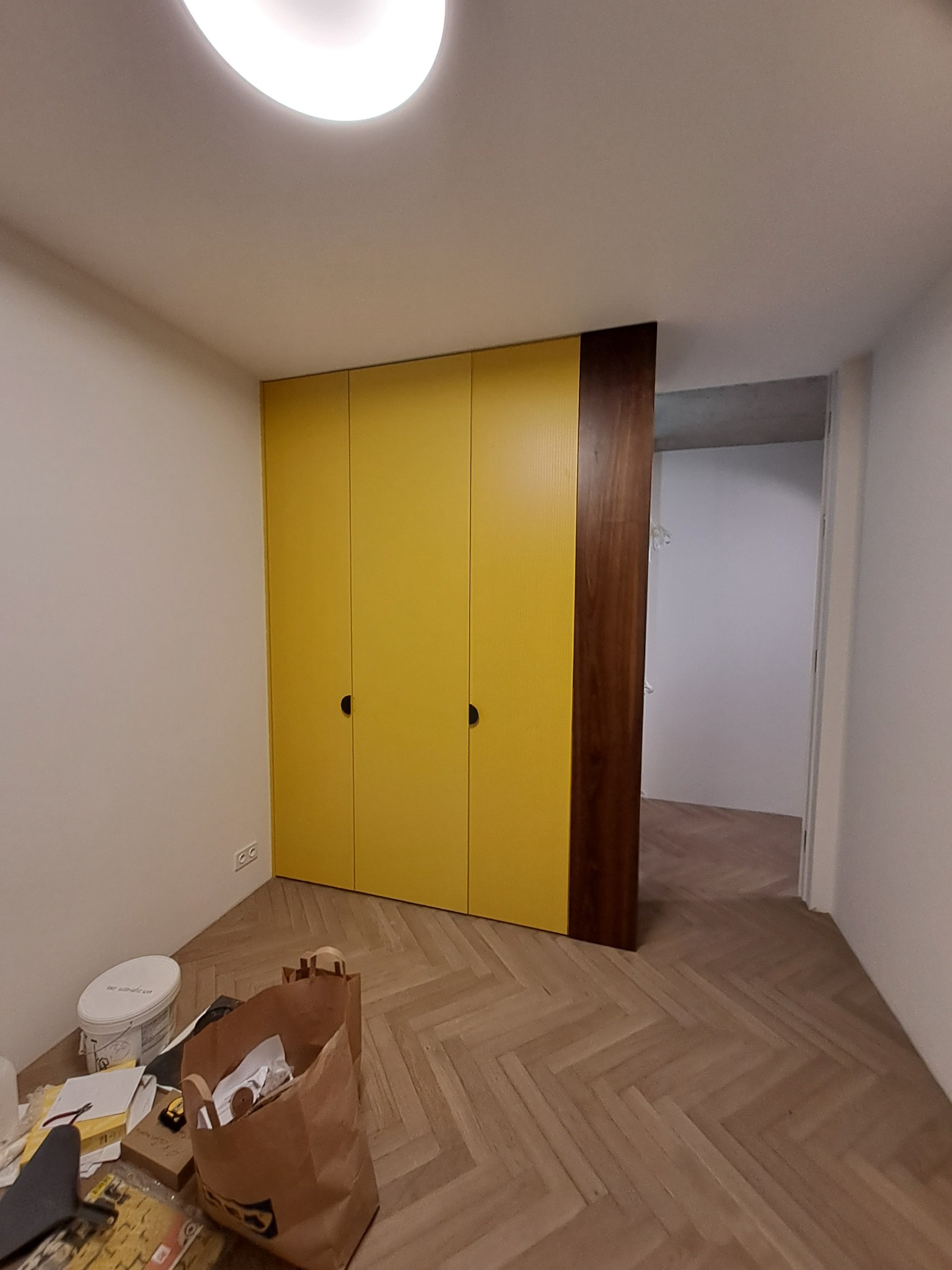 Żółta, trzydrzwiowa szafa z ciemnobrązowym bokiem, na parkiecie ułożonym w jodełkę, widok na wnętrze pomieszczenia, w rogu ślady po remoncie.