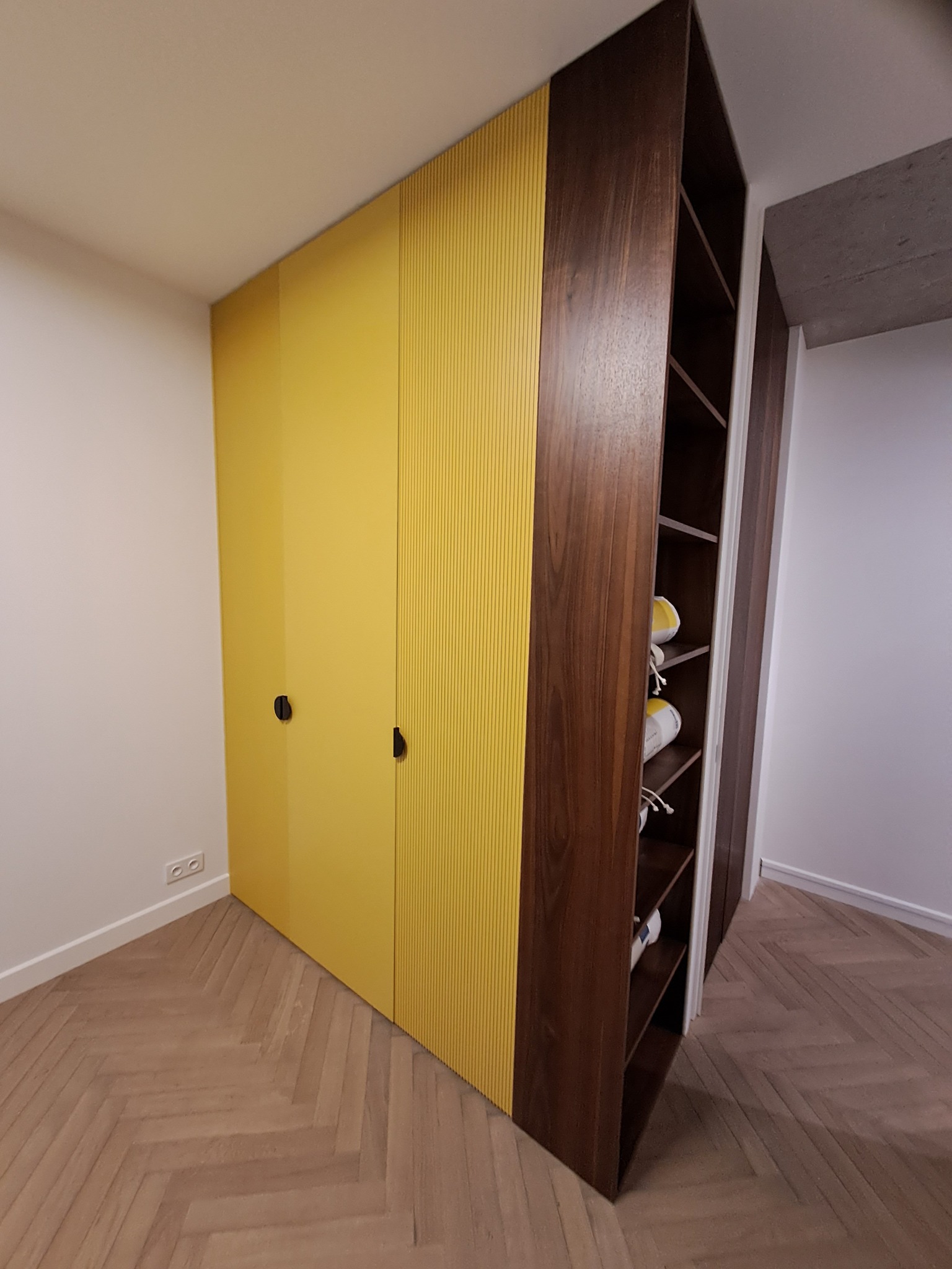 Żółta szafa wnękowa z ryflowanymi frontami i otwartymi półkami z ciemnego drewna, widoczne buty na półkach, parkiet ułożony w jodełkę.