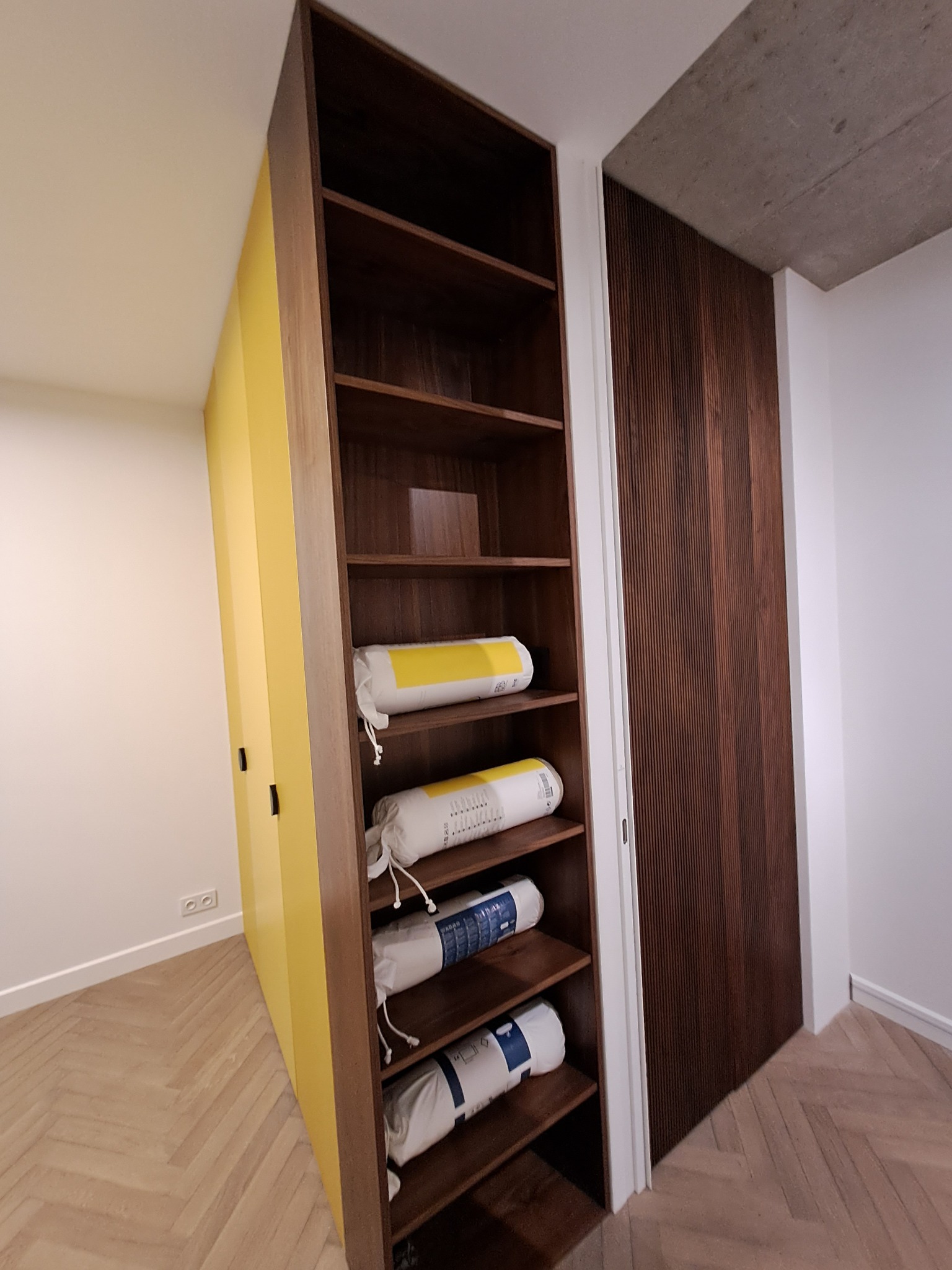Wysoka, brązowa, drewniana półka z żółtymi drzwiczkami obok, wypełniona zwiniętymi materacami w białych pokrowcach, w nowoczesnym wnętrzu z parkietem w jodełkę.