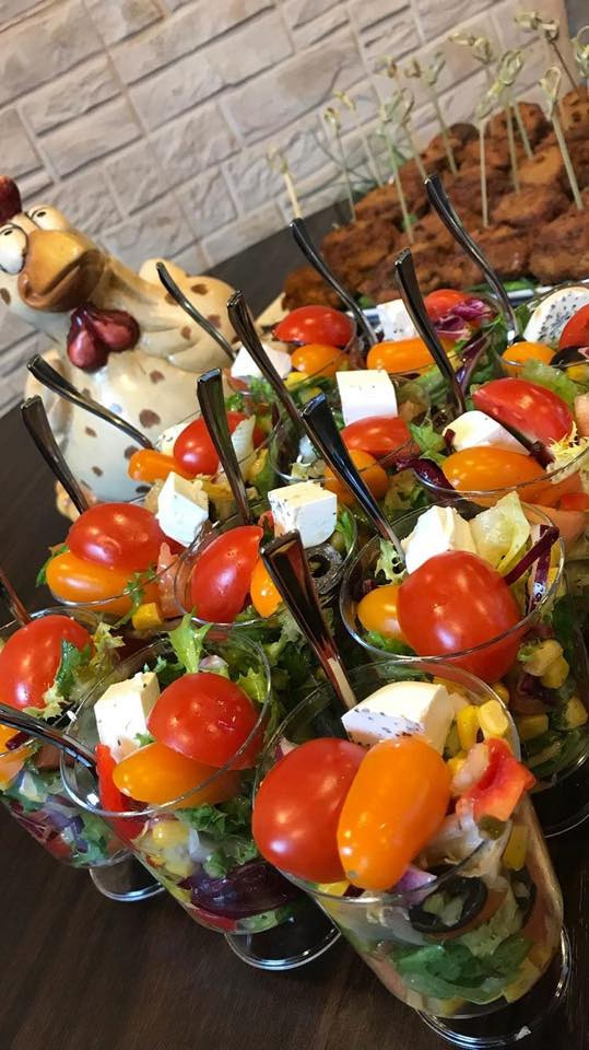 Szklanki z sałatką warzywną, serem feta i pomidorkami koktajlowymi, ułożone rzędami na stole, z dekoracyjną figurką kurczaka w tle.