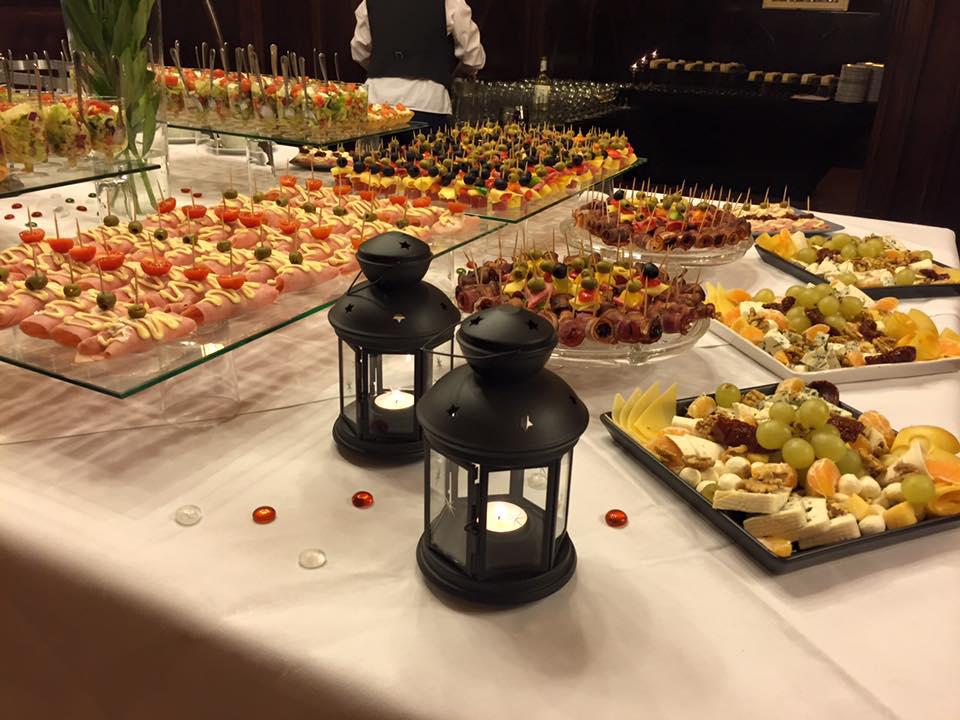 Stół cateringowy z różnorodnymi przekąskami: roladki z szynki z sosem, koreczki z oliwkami i pomidorami, tace z serami, winogronami i owocami, dekorowany lampionami i szklanymi kamyczkami.