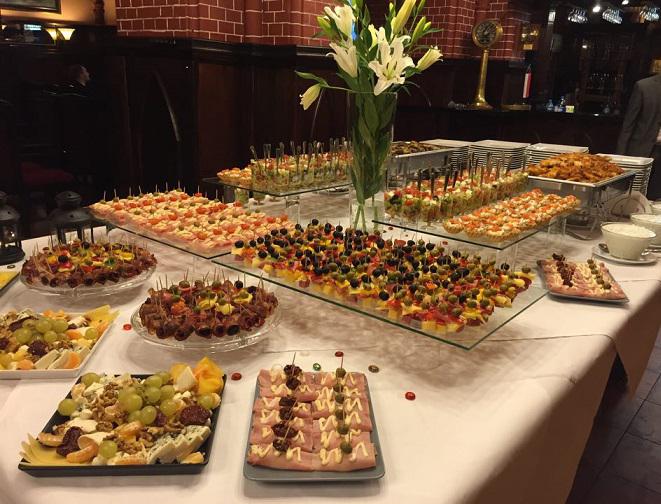 Stół z różnorodnymi przekąskami: koreczki z oliwkami i pomidorkami, roladki z szynki, kompozycje serów i owoców, ustawione na szklanych i porcelanowych naczyniach, w tle wazon z białymi liliami.