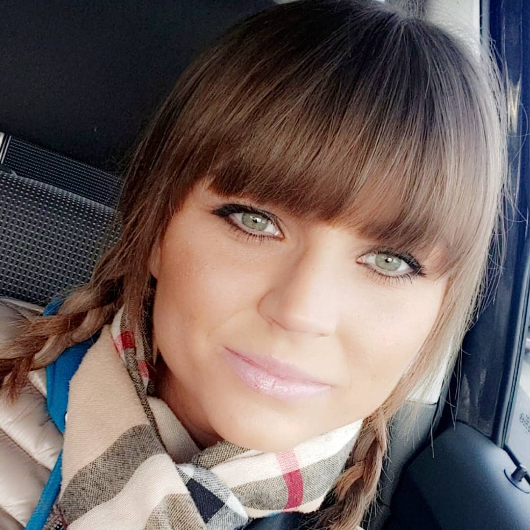 Portret kobiety z zielonymi oczami, w brązowej kurtce i szaliku w kratę, włosy splecione w warkocze, z lekkim uśmiechem, w naturalnym świetle wewnątrz pojazdu.