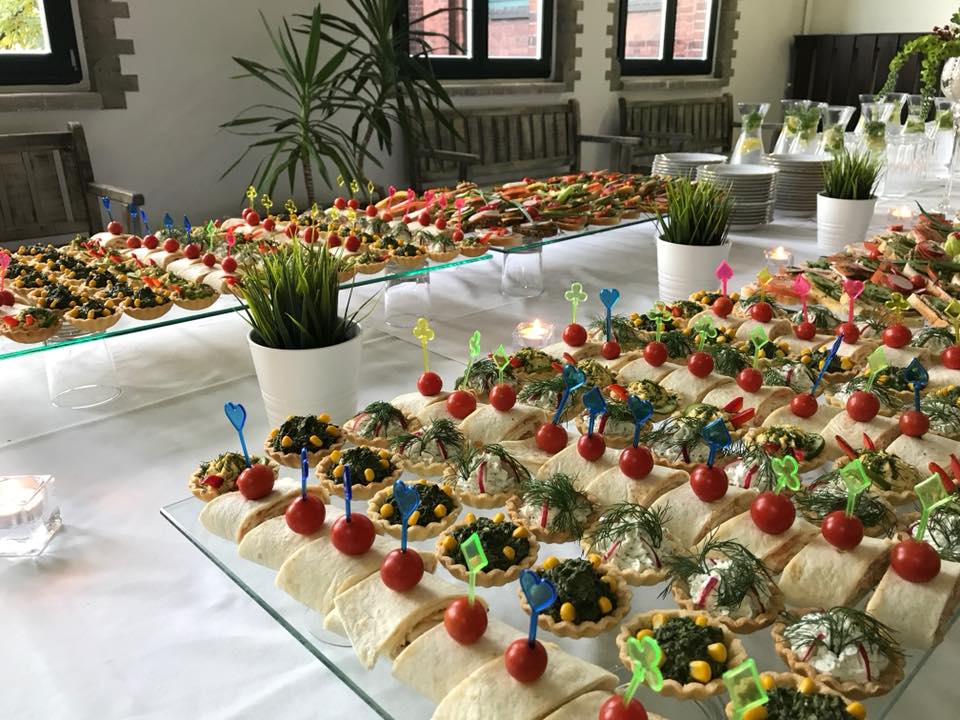 Stół z białym obrusem, na którym znajdują się różnorodne przekąski: tartaletki z farszem warzywnym, roladki z tortilli z pomidorkiem cherry na wykałaczce, kanapki i dekoracyjne rośliny w białych...