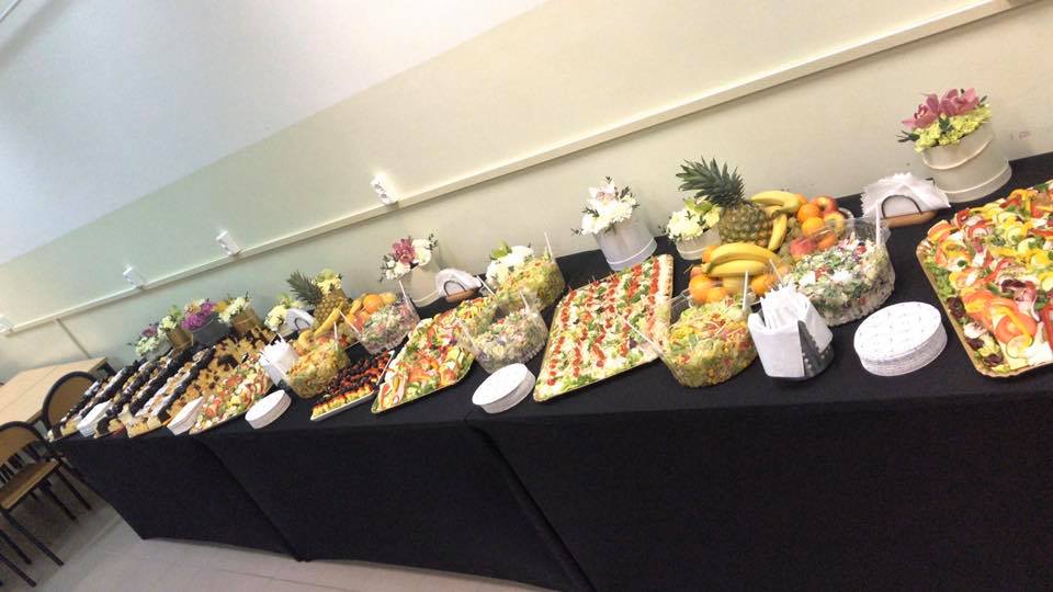 Stół cateringowy z różnorodnymi przekąskami, sałatkami, owocami i dekoracjami kwiatowymi w białych doniczkach, przykryty czarnym obrusem.