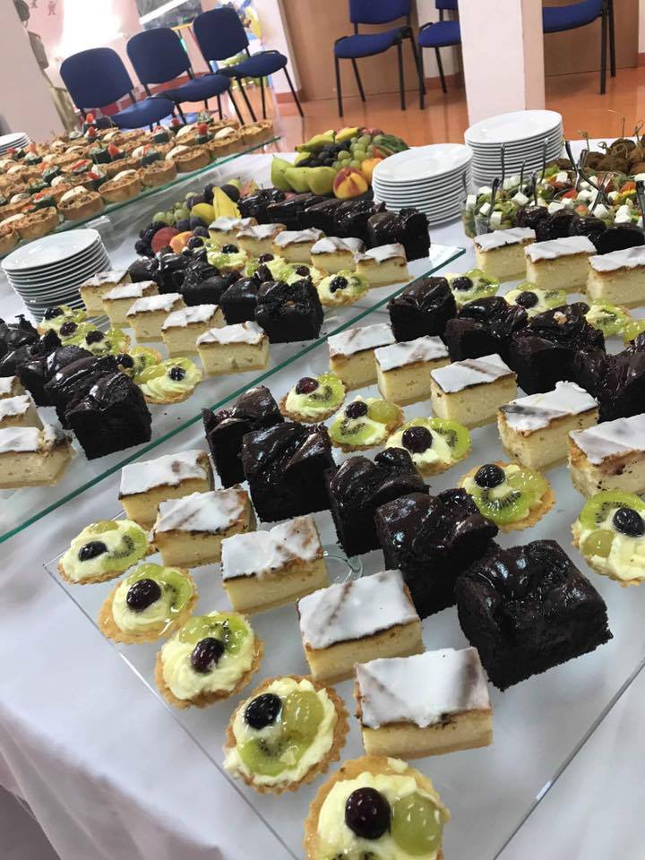 Stół z różnorodnymi mini deserami: tartaletki z owocami, ciasta czekoladowe i biszkoptowe z lukrem, ułożone na szklanej powierzchni, w tle talerze i owoce.