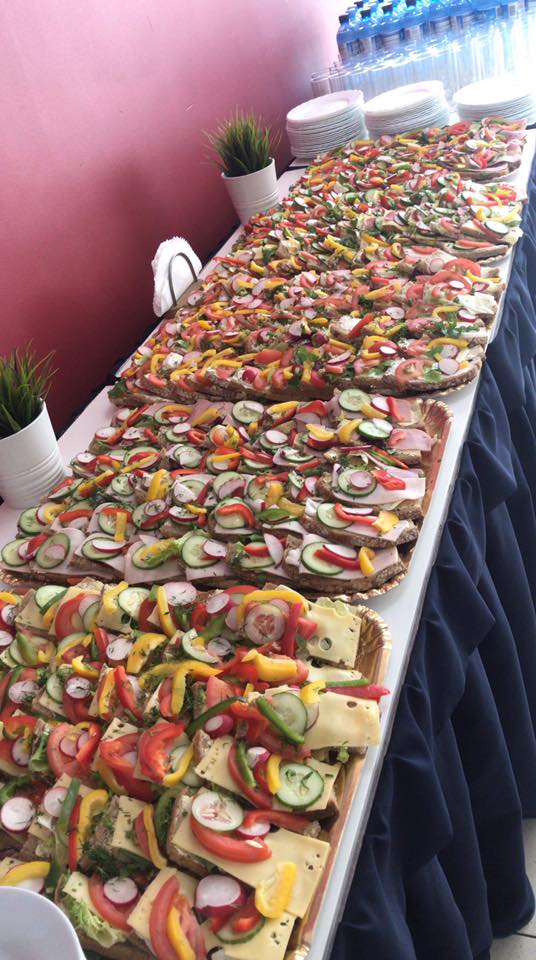 Długi stół z licznymi kanapkami z ciemnego pieczywa, udekorowanymi warzywami: rzodkiewką, ogórkiem, papryką i pomidorami, oraz butelkami wody w tle.