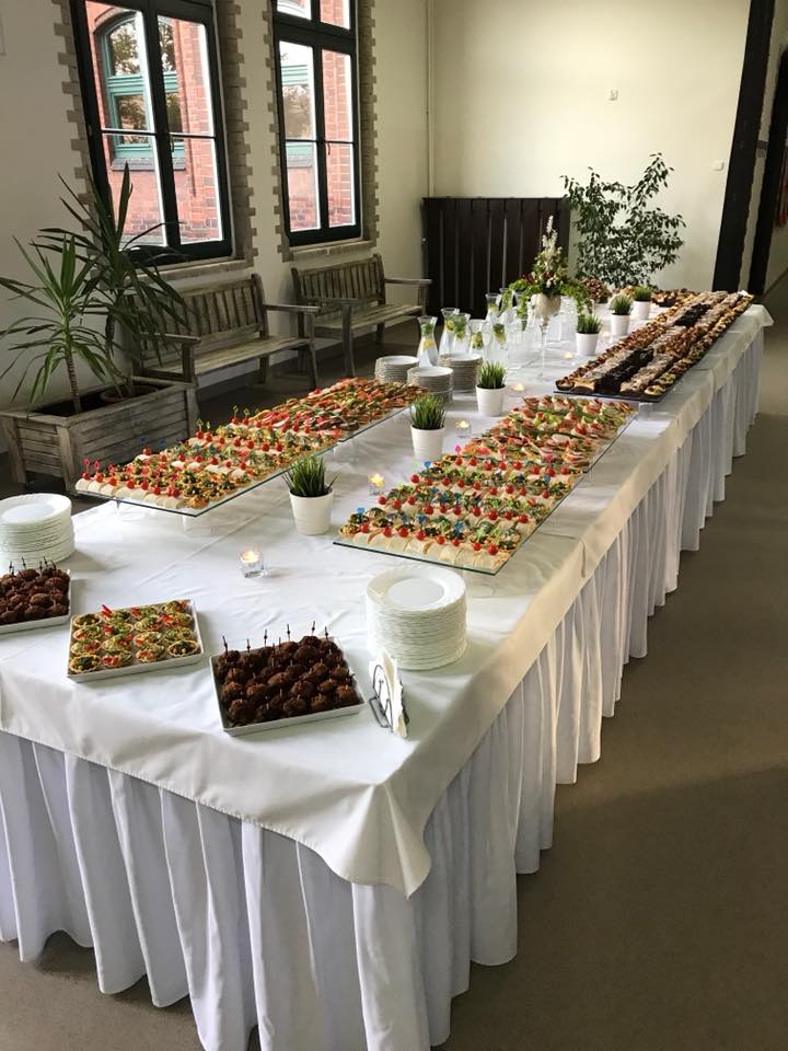 Długi stół udekorowany białym obrusem, zastawiony różnorodnymi przekąskami, mini kanapeczkami, deserami i napojami w szklanych karafkach, przygotowany na przyjęcie w przestronnym wnętrzu z dużymi...