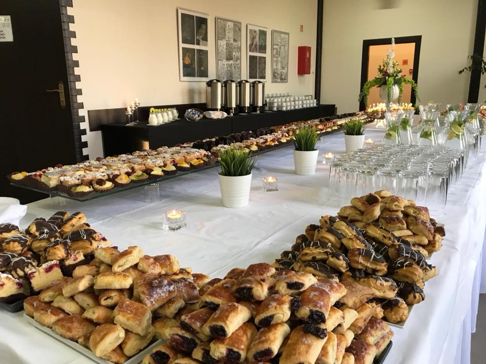 Długi stół z białym obrusem zastawiony różnorodnymi ciastami, babeczkami, ciasteczkami, szklankami z napojami i dekoracjami roślinnymi, przygotowany na przyjęcie.