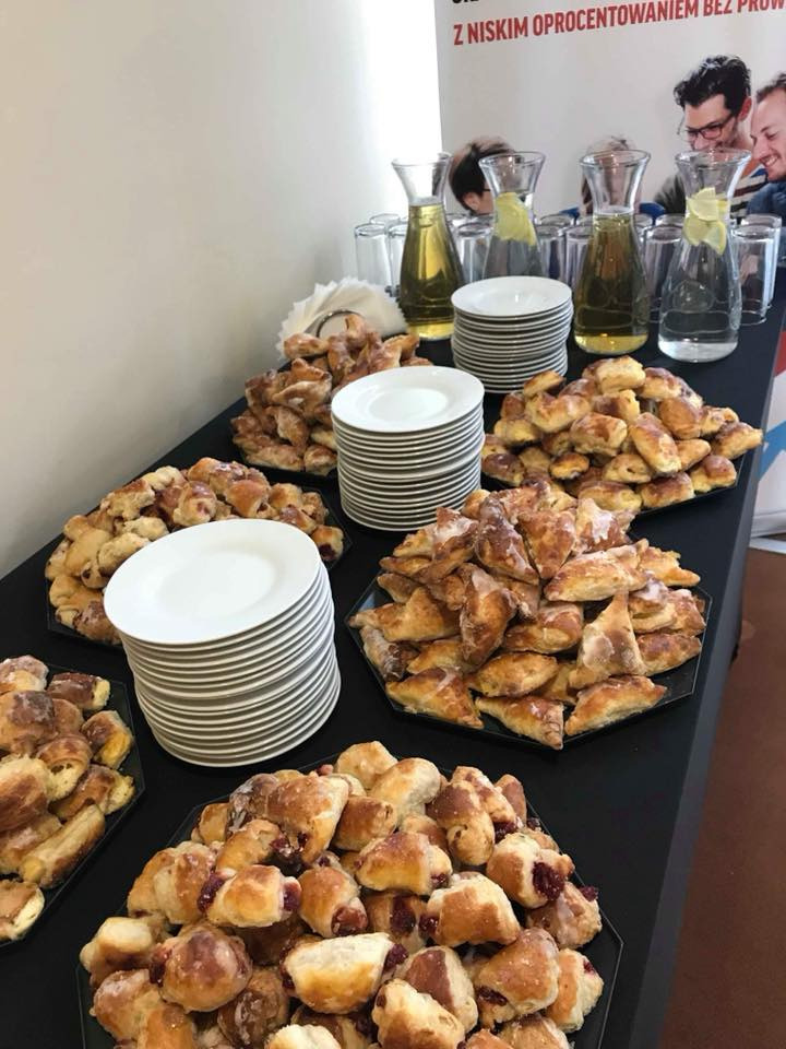 Stół z czarnym obrusem zastawiony talerzami z rogalikami z nadzieniem owocowym, trójkątnymi ciasteczkami oraz dzbankami z lemoniadą, w tle baner reklamowy.