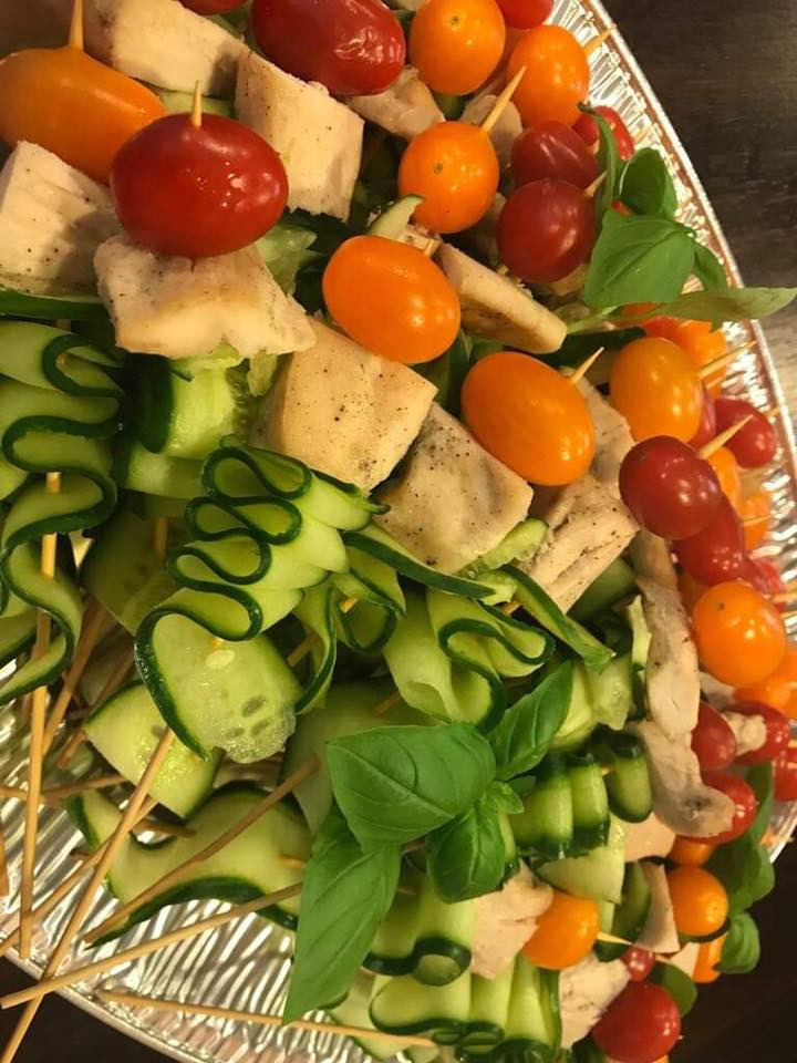 Zbliżenie na srebrną tacę z kolorowymi szaszłykami: spiralne ogórki, pomidorki koktajlowe (czerwone i pomarańczowe), kostki sera oraz świeże listki bazylii, wszystko nabite na drewniane patyczki.