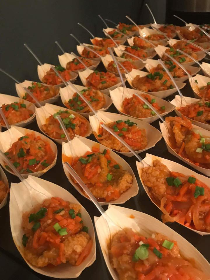 Apetizing finger food: kawałki kurczaka w sosie pomarańczowym, podane w biodegradowalnych łódeczkach z drewna z plastikowymi mini-widelczykami, udekorowane posiekanym szczypiorkiem, ułożone rzędami...