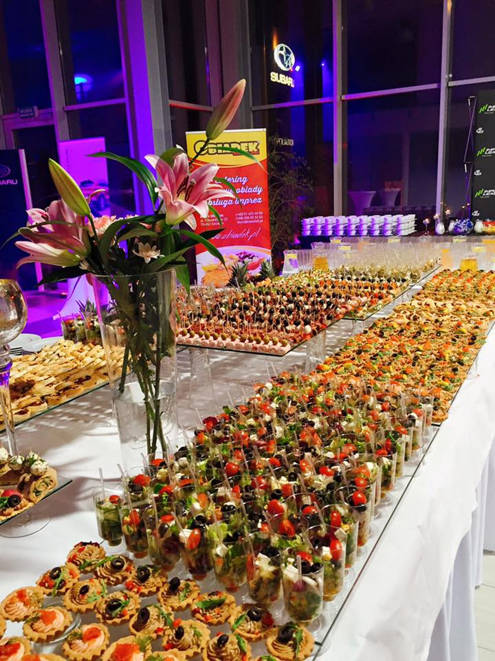 Stół z różnorodnymi przekąskami na przyjęciu, w tle dekoracja kwiatowa i logo Subaru.