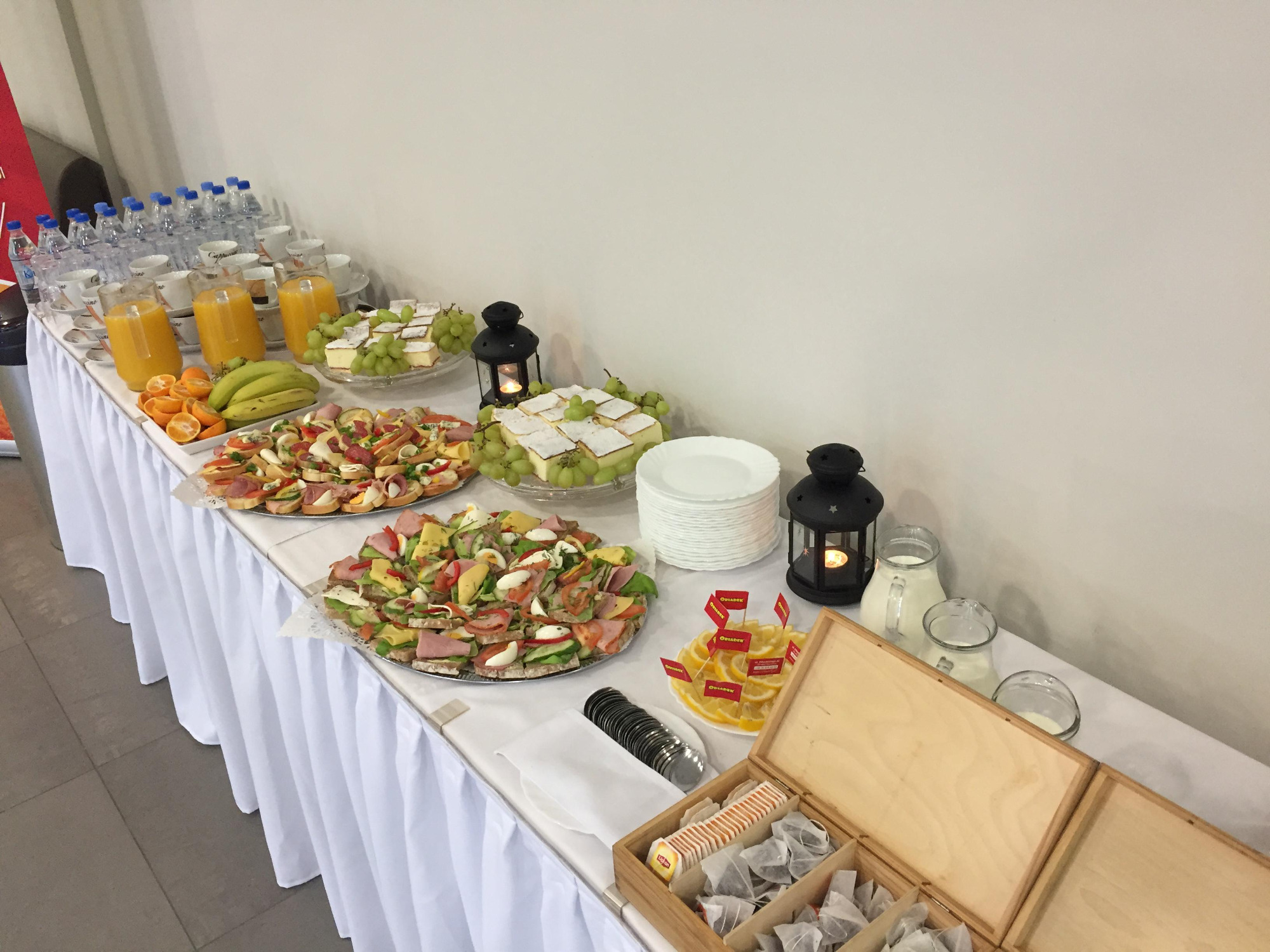 Stół cateringowy z kanapkami, ciastami udekorowanymi winogronami, owocami, dzbankami soku i mleka, wodą butelkowaną, filiżankami oraz drewnianą skrzynką z herbatami, oświetlony dekoracyjnymi...