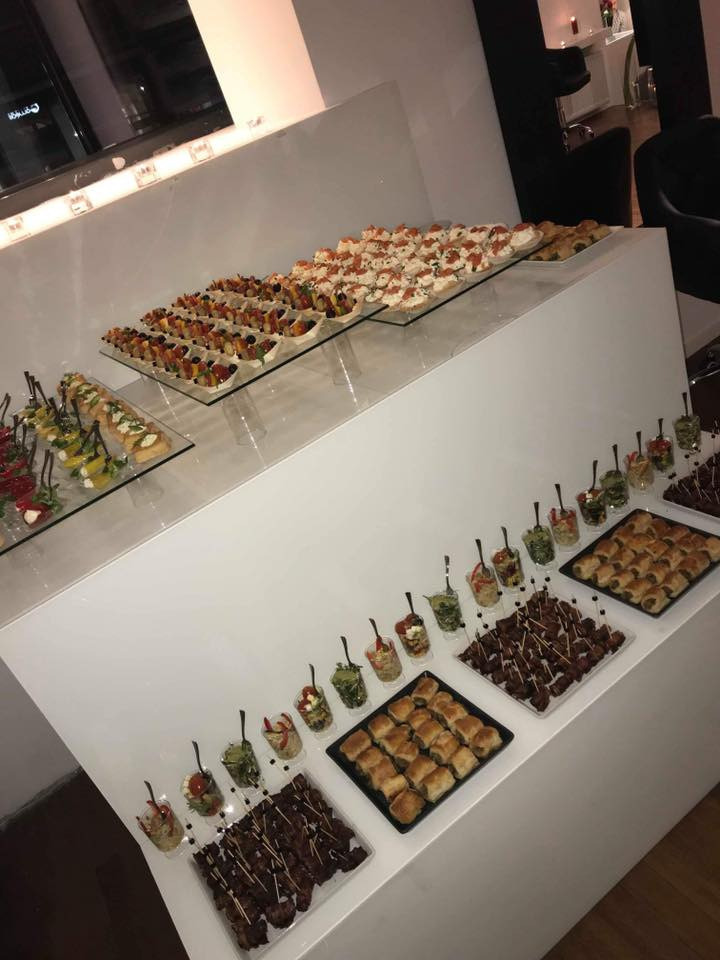 Różnorodne przekąski na białym stole: koreczki, mini sałatki w szklankach, tartinki z pomidorami i owocowe desery w małych pojemnikach.