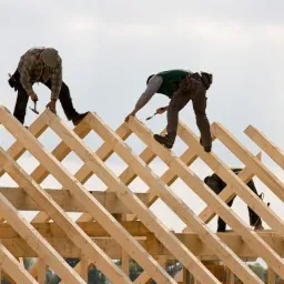 Trzech dekarzy montuje drewnianą konstrukcję dachu na tle jasnego nieba, używając młotków i narzędzi w pasach.