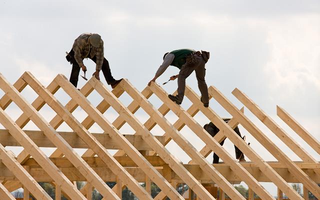 Trzech dekarzy montuje drewnianą konstrukcję dachu na tle jasnego nieba, używając młotków i narzędzi w pasach.