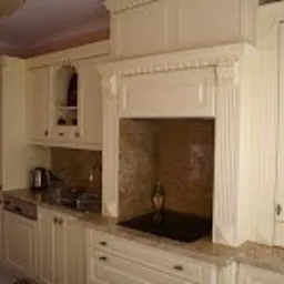 Klasyczna, kremowa zabudowa kuchenna z dekoracyjnym okapem i blatem z jasnego kamienia, widok na fragment ściany z delikatnym wzorem.