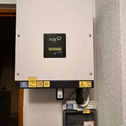 Biała obudowa falownika FOX ESS z wyświetlaczem i naklejkami ostrzegawczymi, zamontowana na ścianie o chropowatej fakturze, podłączona do licznika energii elektrycznej z oznaczeniem PV.