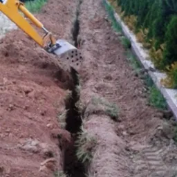 Żółta koparka wykonuje wąski wykop w ziemi, widoczne ślady opon, w tle rząd tuj.