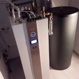 Nowoczesna instalacja grzewcza z białym panelem sterowania, zbiornikiem akumulacyjnym i licznymi rurami, widoczna w pomieszczeniu technicznym z jasnymi płytkami podłogowymi.