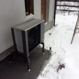 Zewnętrzna jednostka pompy ciepła powietrze-woda, zamontowana przy ścianie budynku pokrytego białym tynkiem, z widocznym oknem i fragmentem elewacji w zimowej scenerii.