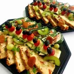 Dwie czarne tace z elegancko ułożonymi przekąskami: pasztetami z sezamem, roladkami szpinakowymi, pomidorkami koktajlowymi, oliwkami, kiwi, winogronami i morelami na wykałaczkach.