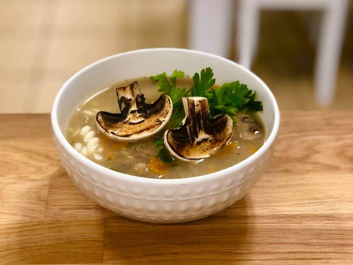 Mushroom soup in a white textured bowl, garnished with grilled mushroom slices and parsley, set on a light wood surface with a blurred white chair in the background.