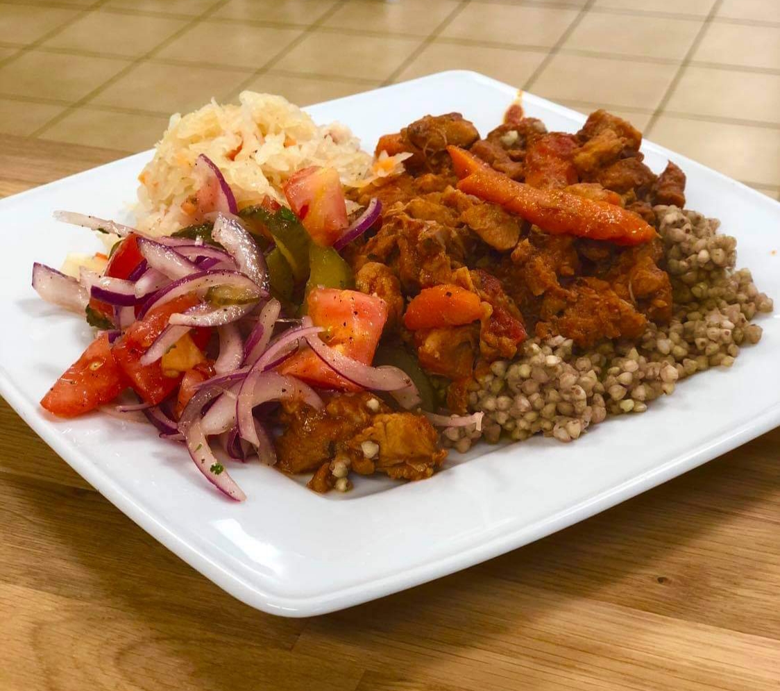 Danie na białym kwadratowym talerzu: kasza gryczana, gulasz warzywny z mięsem, surówka z kapusty kiszonej i sałatka z pomidorów i czerwonej cebuli.