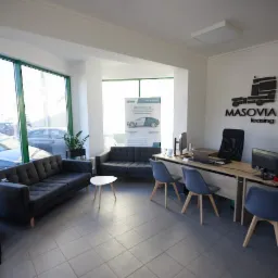 Jasne biuro z szarymi sofami dla klientów, biurkami z komputerami i logo firmy na ścianie, widok na samochody za oknem.