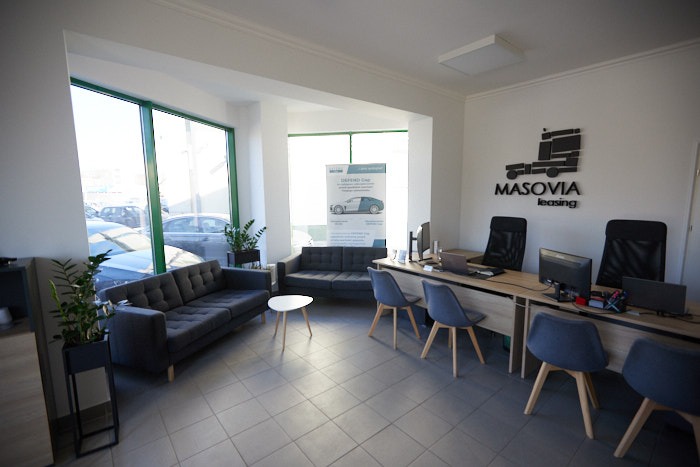 Jasne biuro z szarymi sofami dla klientów, biurkami z komputerami i logo firmy na ścianie, widok na samochody za oknem.