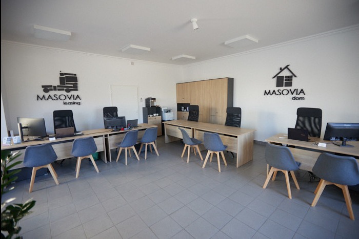 Jasne biuro firmy Masovia z logo leasingu ciężarówek i domów na ścianie, wyposażone w drewniane biurka, szare krzesła, komputery i szafę w tle.