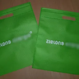 Dwie zielone, materiałowe torby z wyciętymi uchwytami i białym nadrukiem 'zielona' oraz zamazanym logo, leżące na drewnianym podłożu.
