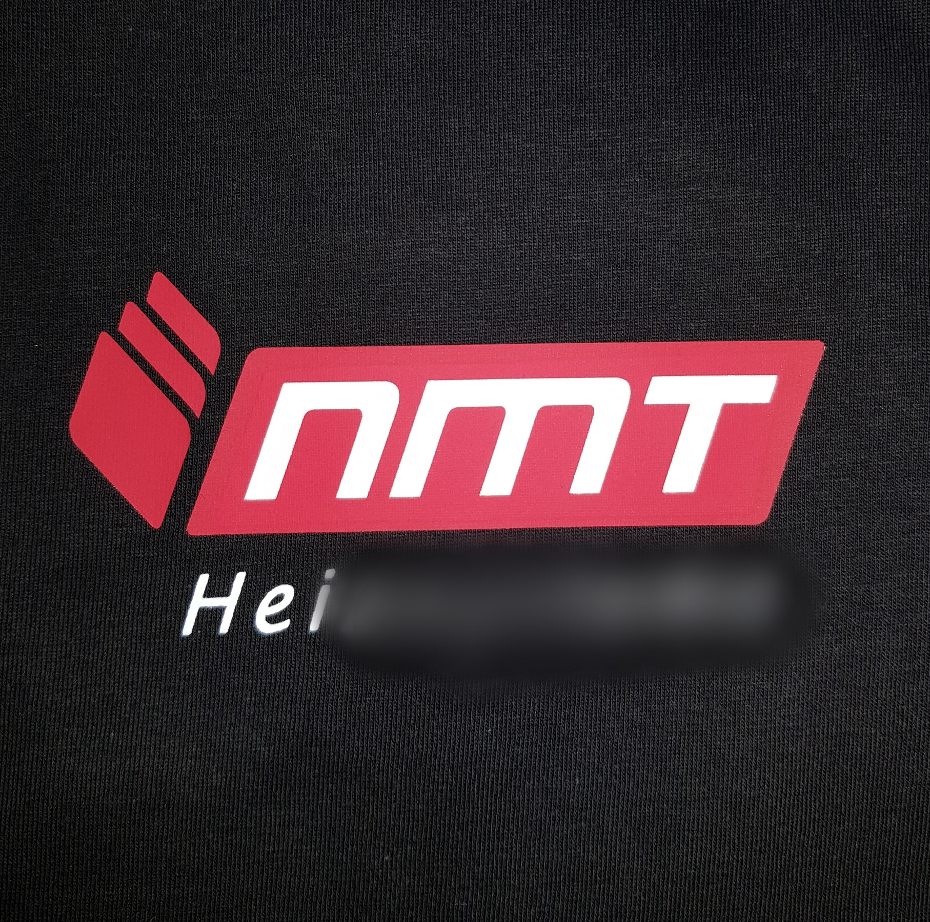 Czerwono-białe logo 'NMT' na czarnej koszulce z widoczną strukturą materiału; pod logiem częściowo zamazany napis.