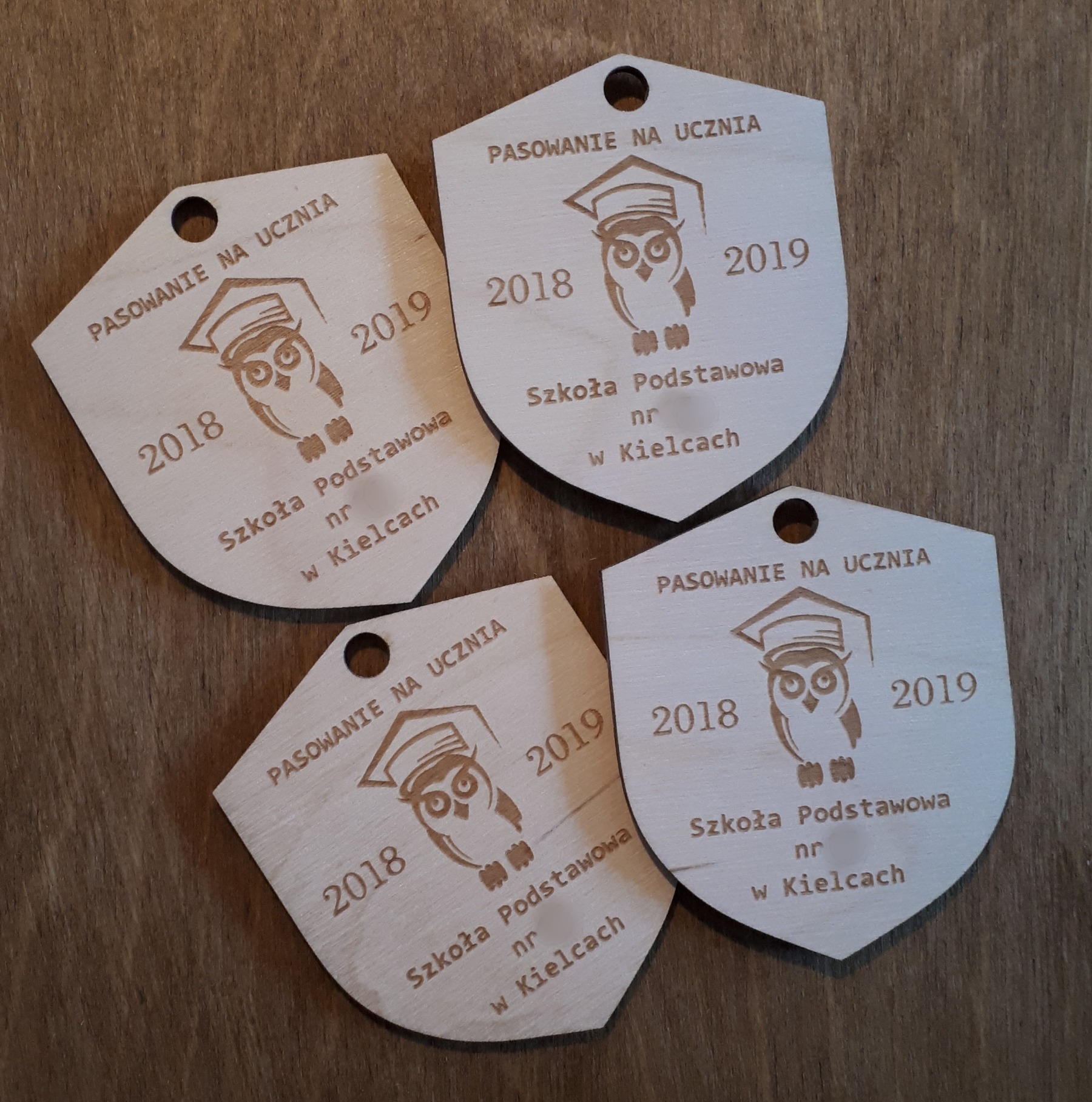 Cztery drewniane zawieszki w kształcie tarczy z wygrawerowanym napisem 'Pasowanie na Ucznia', datami 2018 i 2019, grafiką sowy w birecie oraz nazwą Szkoły Podstawowej nr w Kielcach, leżące...