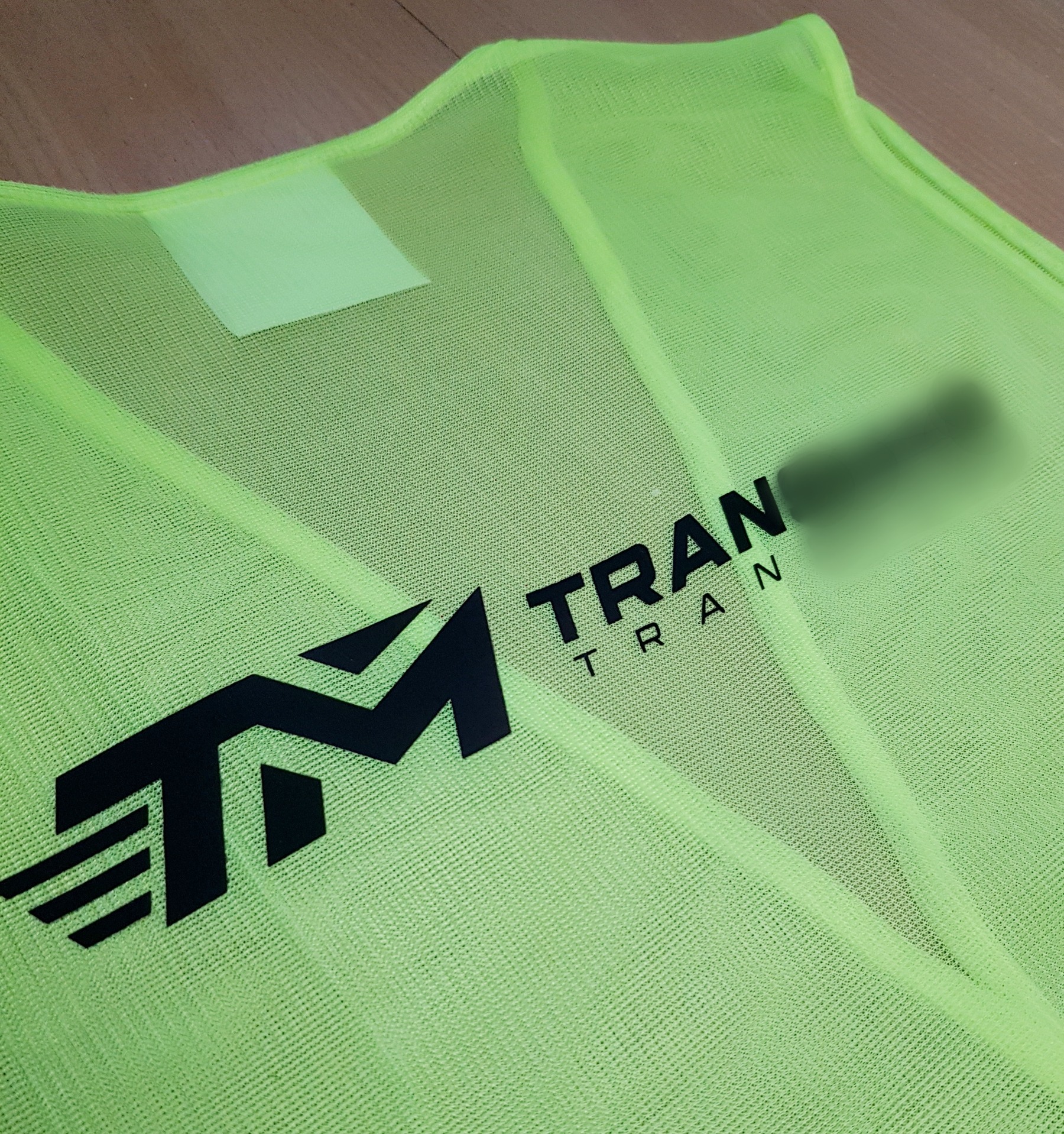 Fluorescencyjna, siateczkowa kamizelka z czarnym nadrukiem logo firmy z Kielc, leżąca na drewnianym podłożu.